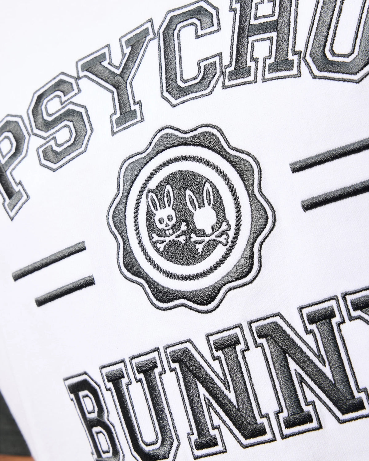 A close-up of the Psycho Bunny Mens London Raglan Graphic Tee (B6U896D200) in white Pima Cotton, featuring the brand name "PSYCHO BUNNY" in bold black letters. Centrally placed, there is a varsity-style graphic emblem with two stylized bunny heads and crossed bones, highlighting a heavyweight design in black and white.