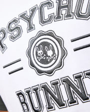 A close-up of the Psycho Bunny Mens London Raglan Graphic Tee (B6U896D200) in white Pima Cotton, featuring the brand name "PSYCHO BUNNY" in bold black letters. Centrally placed, there is a varsity-style graphic emblem with two stylized bunny heads and crossed bones, highlighting a heavyweight design in black and white.