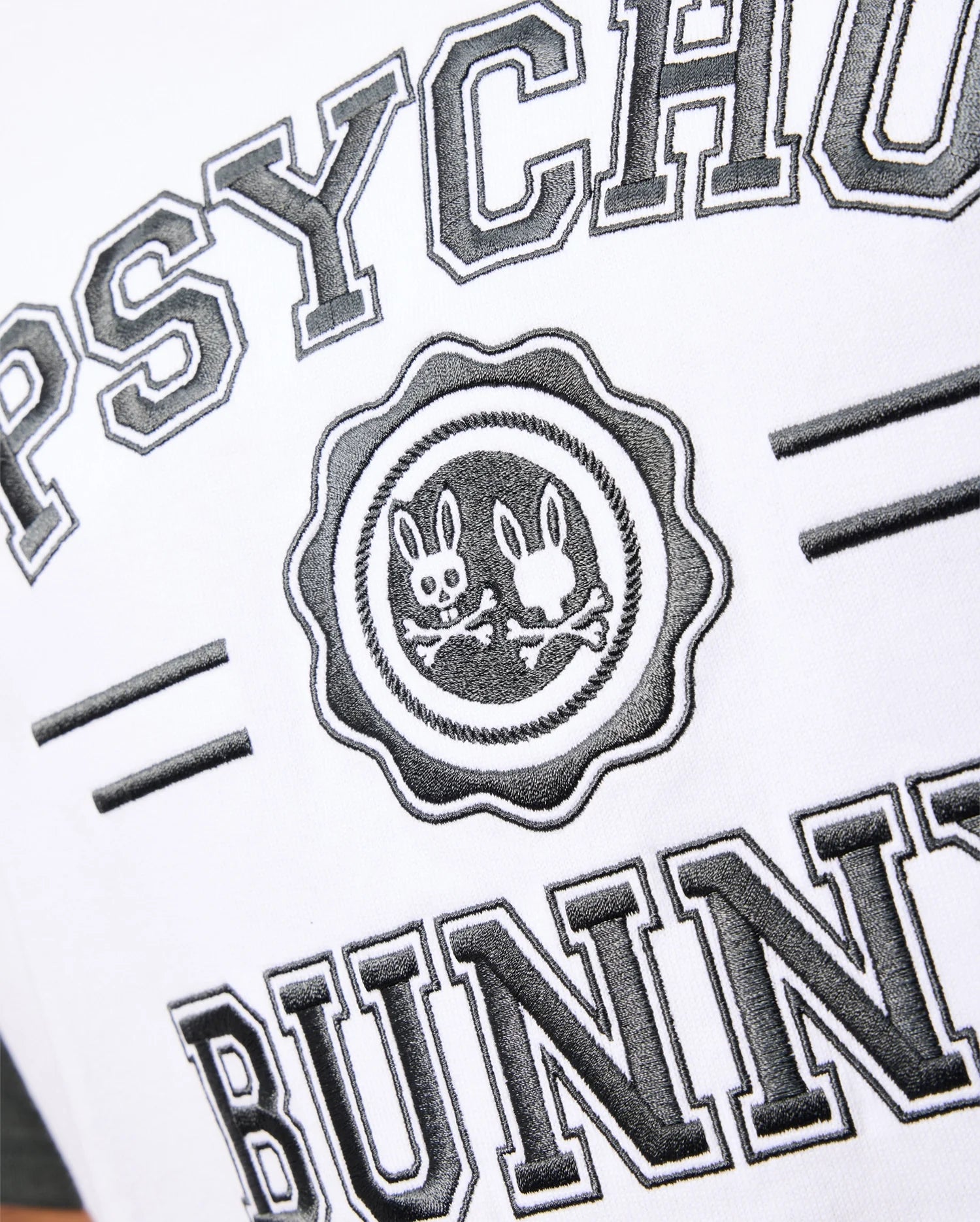 A close-up of the Psycho Bunny Mens London Raglan Graphic Tee (B6U896D200) in white Pima Cotton, featuring the brand name "PSYCHO BUNNY" in bold black letters. Centrally placed, there is a varsity-style graphic emblem with two stylized bunny heads and crossed bones, highlighting a heavyweight design in black and white.