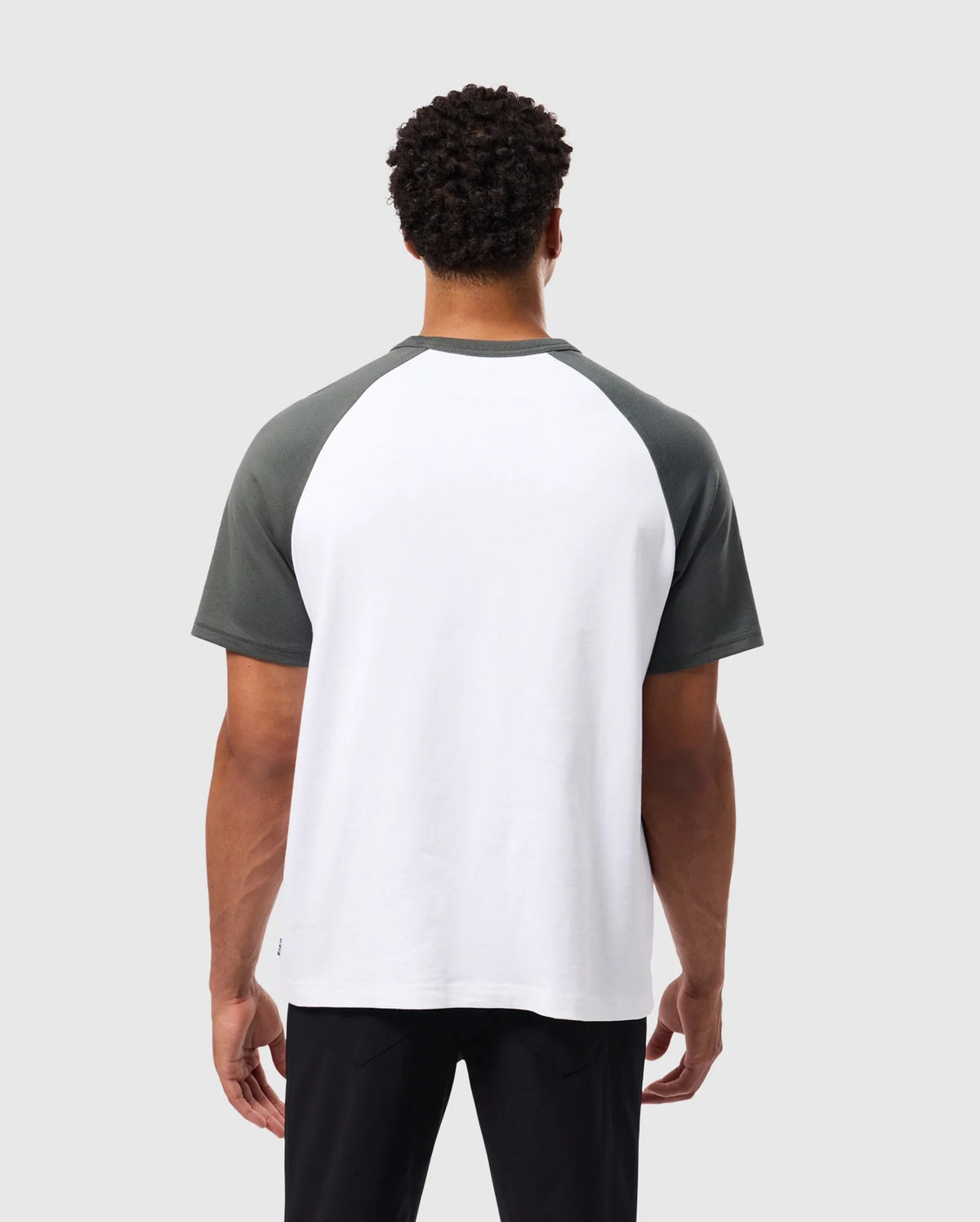A person with short curly hair is wearing a Psycho Bunny MENS LONDON RAGLAN GRAPHIC TEE - B6U896D200, featuring dark green sleeves and a white body. They are facing away from the camera against a plain gray background, highlighting the versatility of this varsity graphic style.