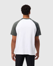 A person with short curly hair is wearing a Psycho Bunny MENS LONDON RAGLAN GRAPHIC TEE - B6U896D200, featuring dark green sleeves and a white body. They are facing away from the camera against a plain gray background, highlighting the versatility of this varsity graphic style.