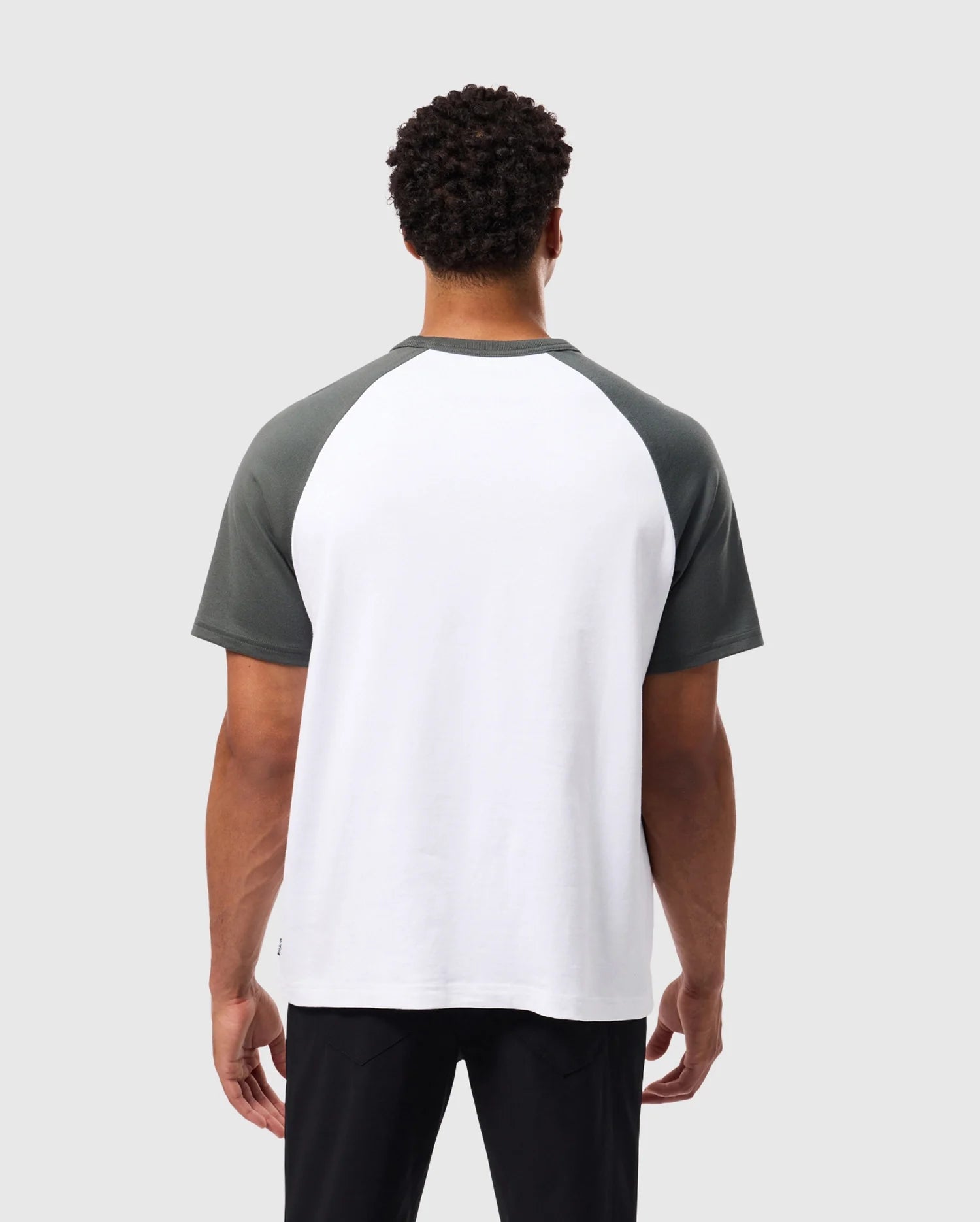 A person with short curly hair is wearing a Psycho Bunny MENS LONDON RAGLAN GRAPHIC TEE - B6U896D200, featuring dark green sleeves and a white body. They are facing away from the camera against a plain gray background, highlighting the versatility of this varsity graphic style.
