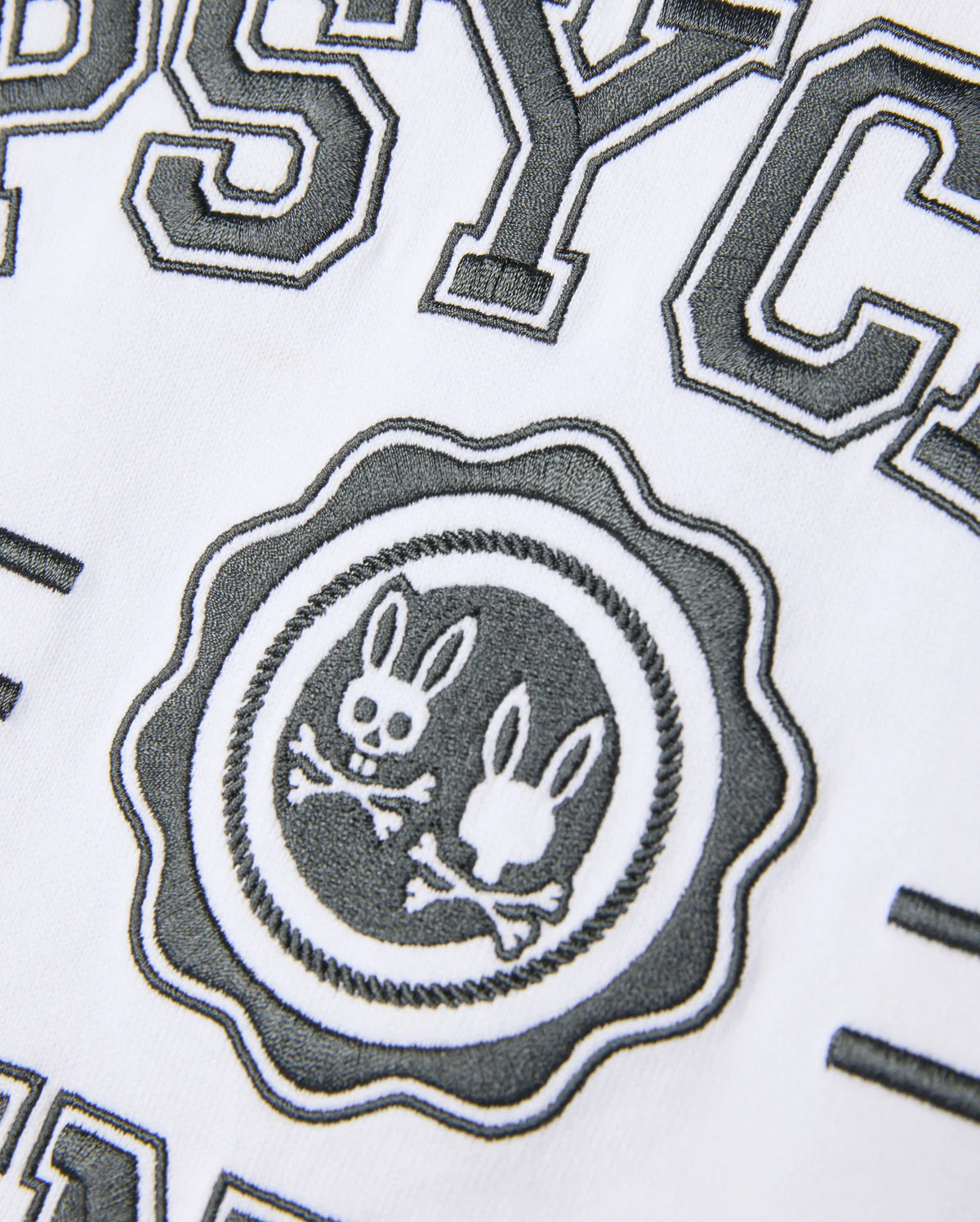 Close-up of a white Psycho Bunny Pima Cotton fabric with black, textured embroidery. This Heavy Weight Mens London Raglan Graphic Tee - B6U896D200 showcases the word "PSYC" above a circular logo featuring two bunny heads and crossbones, accompanied by partially visible surrounding text.