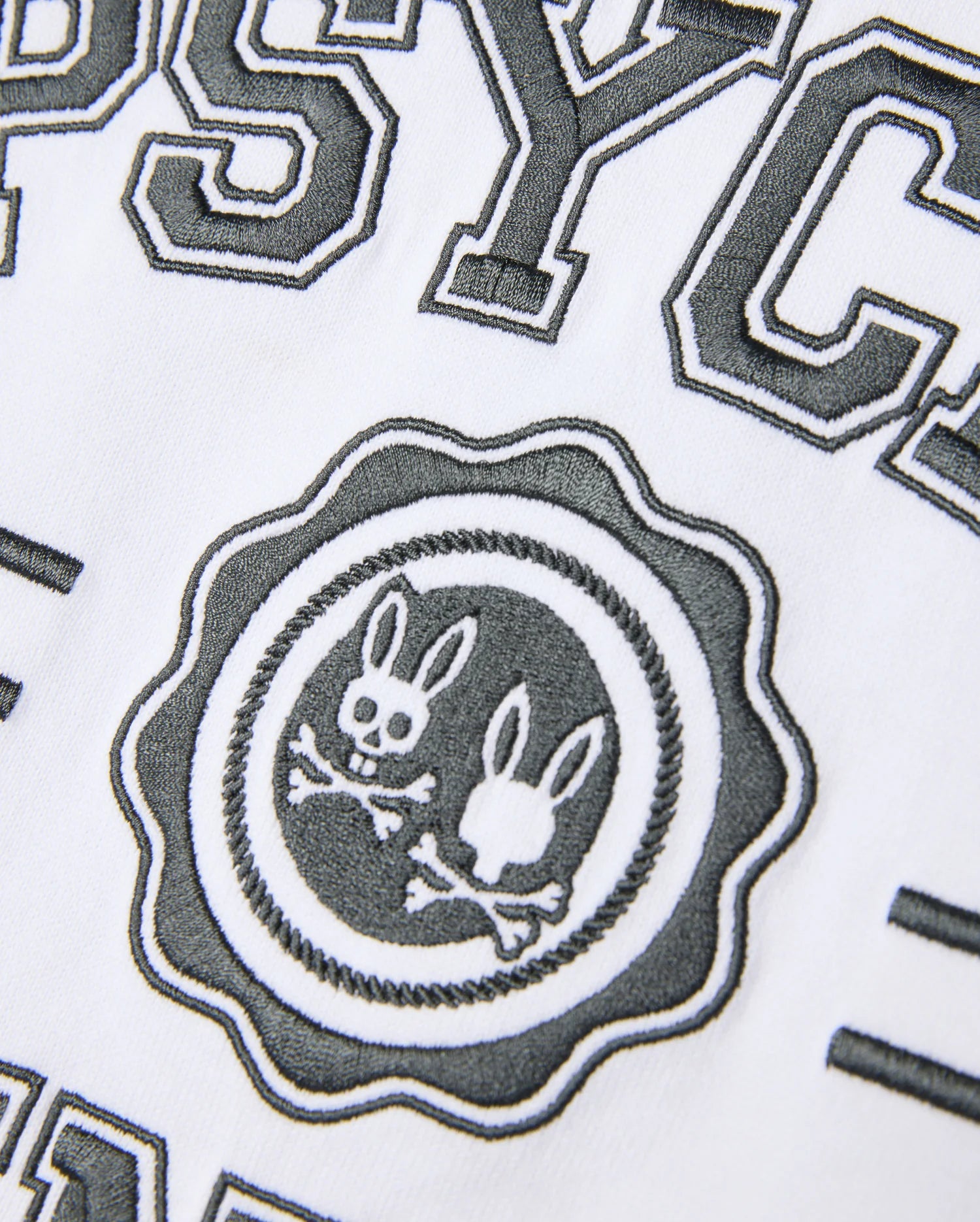Close-up of a white Psycho Bunny Pima Cotton fabric with black, textured embroidery. This Heavy Weight Mens London Raglan Graphic Tee - B6U896D200 showcases the word "PSYC" above a circular logo featuring two bunny heads and crossbones, accompanied by partially visible surrounding text.