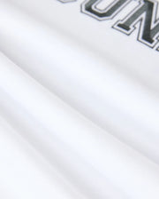 Close-up of white fabric from the Psycho Bunny MENS LONDON RAGLAN GRAPHIC TEE - B6U896D200, featuring black text with the visible letters "OUN" and a partial "R" at the top. The smooth pima cotton fabric is slightly folded, forming soft shadows.