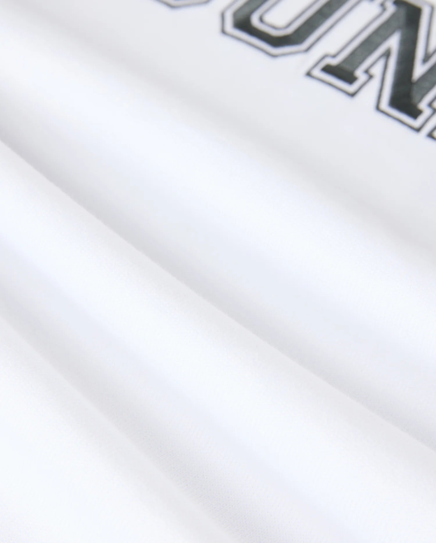 Close-up of white fabric from the Psycho Bunny MENS LONDON RAGLAN GRAPHIC TEE - B6U896D200, featuring black text with the visible letters "OUN" and a partial "R" at the top. The smooth pima cotton fabric is slightly folded, forming soft shadows.
