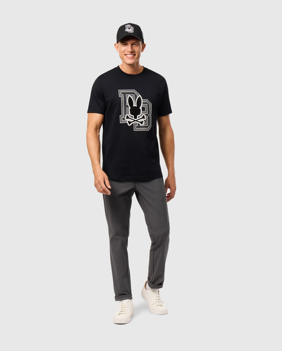 A man is smiling and standing against a plain light gray background, wearing a black cap, a Psycho Bunny MENS ZACK GRAPHIC TEE - B6U897D200 featuring a large stylized Mickey Mouse design, gray pants, and white sneakers. His hands are relaxed by his sides.