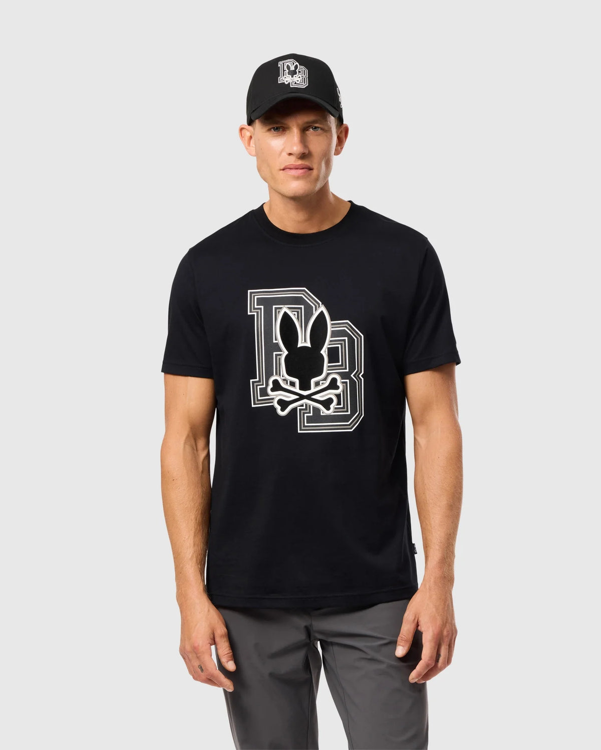 A person wearing a black cap and the Psycho Bunny MENS ZACK GRAPHIC TEE - B6U897D200, featuring a large "DB" logo and a bunny with crossed bones. The individual is standing against a plain light gray background, also dressed in gray pants.
