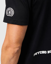 Close-up of a black MENS ZACK GRAPHIC TEE - B6U897D200 from Psycho Bunny, made from soft Pima cotton, with a patch on the sleeve featuring the words "Psycho Bunny" around a bunny skull and crossbones logo. The word "Psycho Bunny" is also partially visible on the back of the T-shirt. The background is plain and grey.