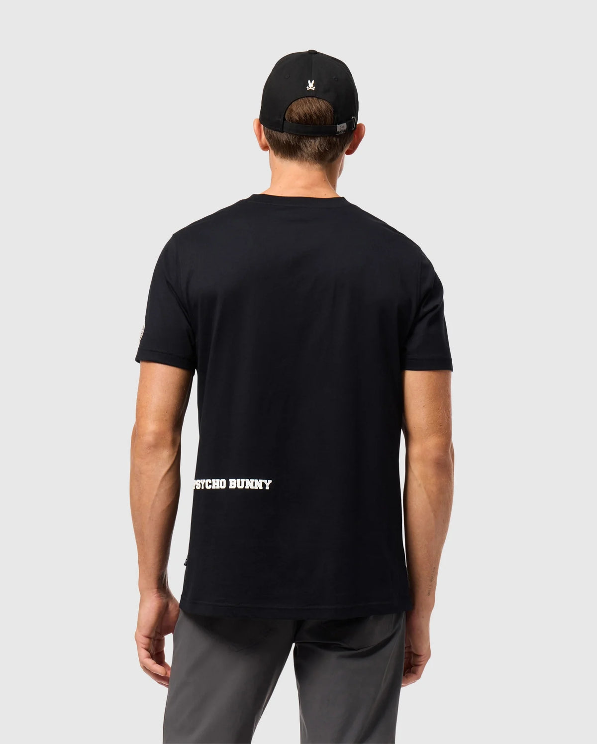 A person wearing a black cap and the Psycho Bunny MENS ZACK GRAPHIC TEE - B6U897D200, featuring "PSYCHO BUNNY" in white near the lower back, is facing away from the camera. The Pima cotton tee is paired with gray pants, set against a plain light gray background.