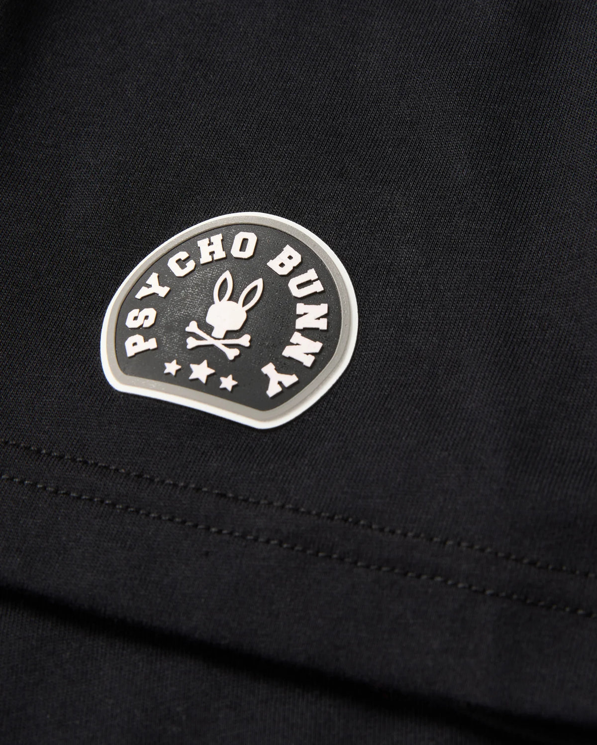 Close-up of the MENS ZACK GRAPHIC TEE - B6U897D200, a black Pima cotton graphic tee from Psycho Bunny. The tee features a distinctive Psycho Bunny logo consisting of a bunny with crossed bones beneath, encircled by the brand name "Psycho Bunny" in curved text. Three stars are also visible beneath the bunny graphic.