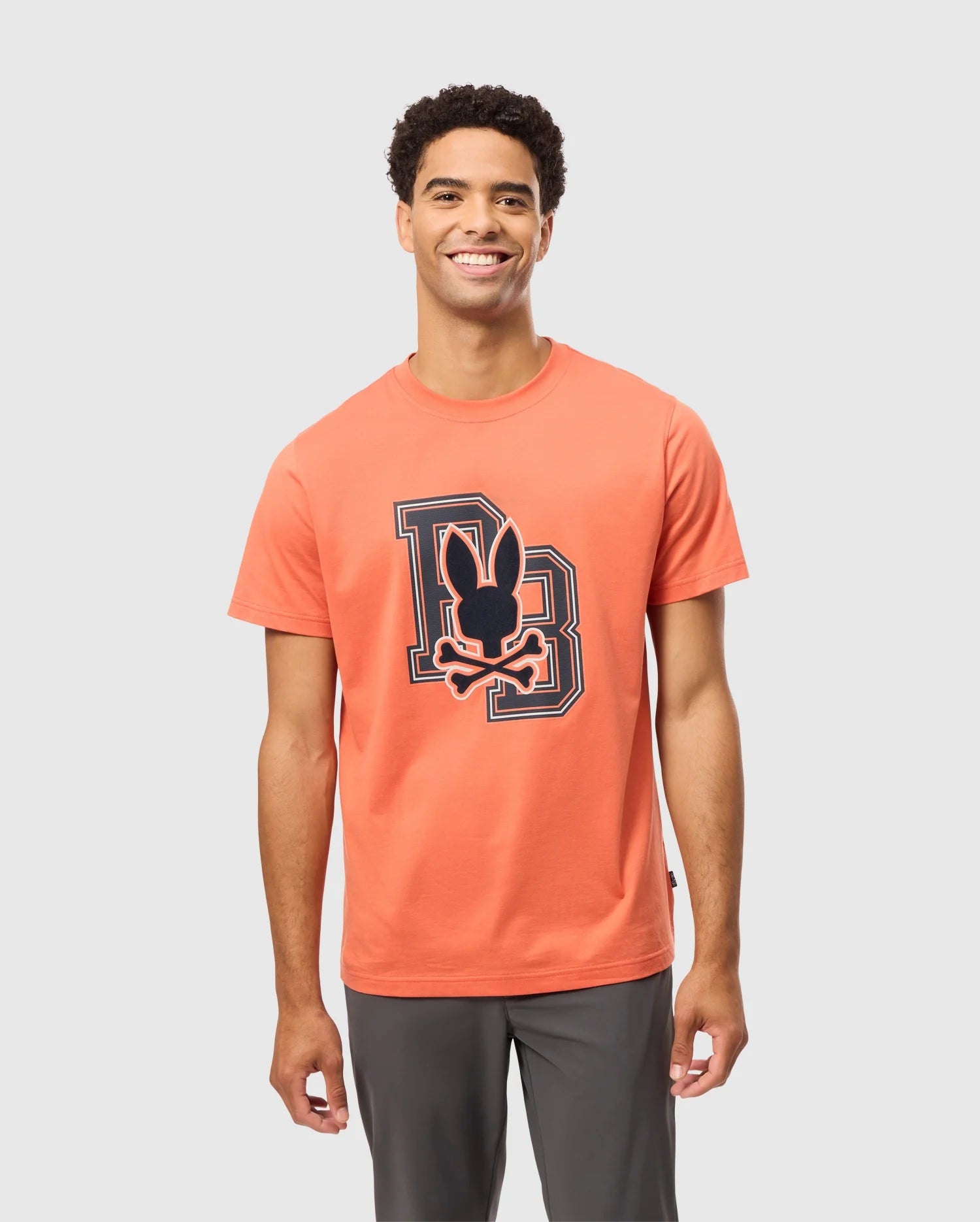 A smiling person models the vibrant orange MENS ZACK GRAPHIC TEE - B6U897D200 made from Pima cotton, showcasing a cartoon character and stylized initials. With short, curly hair, they exude confident charm against a plain light gray background, embodying the bold spirit of Psycho Bunny.