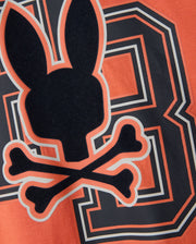 The Psycho Bunny MENS ZACK GRAPHIC TEE - B6U897D200 features an edgy design with a black silhouette of a bunny head and crossed bones, centered over a large "8" on an orange background. This striking tee, made from soft Pima cotton, highlights the bunny and bones outlined in vivid orange and white borders.