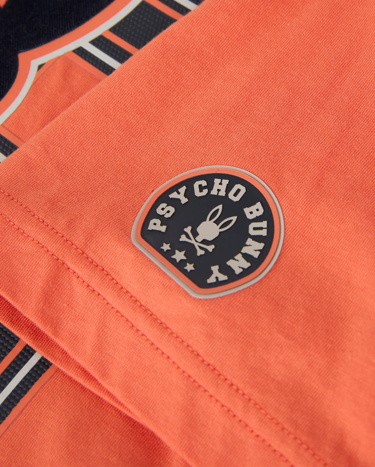 A detailed view of an orange Pima cotton MENS ZACK GRAPHIC TEE by Psycho Bunny, featuring a circular patch with the words "Psycho Bunny" and a bunny emblem wearing a bow tie, encircled by stars.