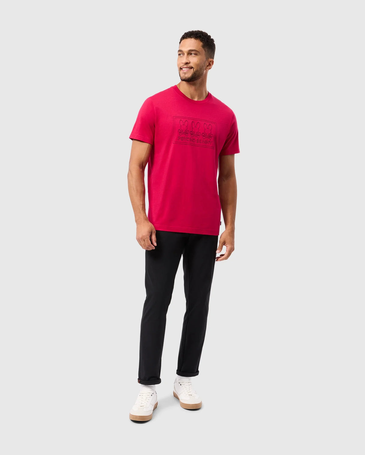 A man with short hair stands smiling, dressed in a bright red Psycho Bunny MENS TACOMA GRAPHIC TEE - B6U909D200 crafted from soft Pima cotton. He pairs it with black pants and white sneakers, his body angled slightly to the side against a plain gray background, with his hands resting naturally by his sides.