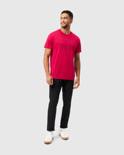 A man with short hair stands smiling, dressed in a bright red Psycho Bunny MENS TACOMA GRAPHIC TEE - B6U909D200 crafted from soft Pima cotton. He pairs it with black pants and white sneakers, his body angled slightly to the side against a plain gray background, with his hands resting naturally by his sides.