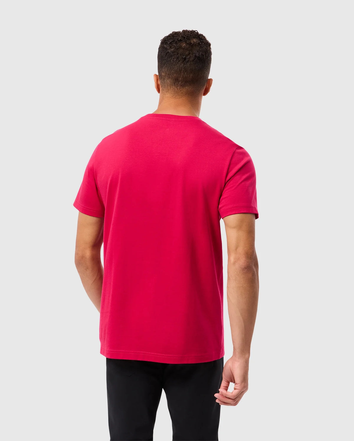 A person with short curly hair is standing with their back to the camera, wearing a vibrant red Psycho Bunny MENS TACOMA GRAPHIC TEE - B6U909D200 and black pants. The fabric of the shirt looks soft, possibly Pima cotton. The background is a plain, light grey.