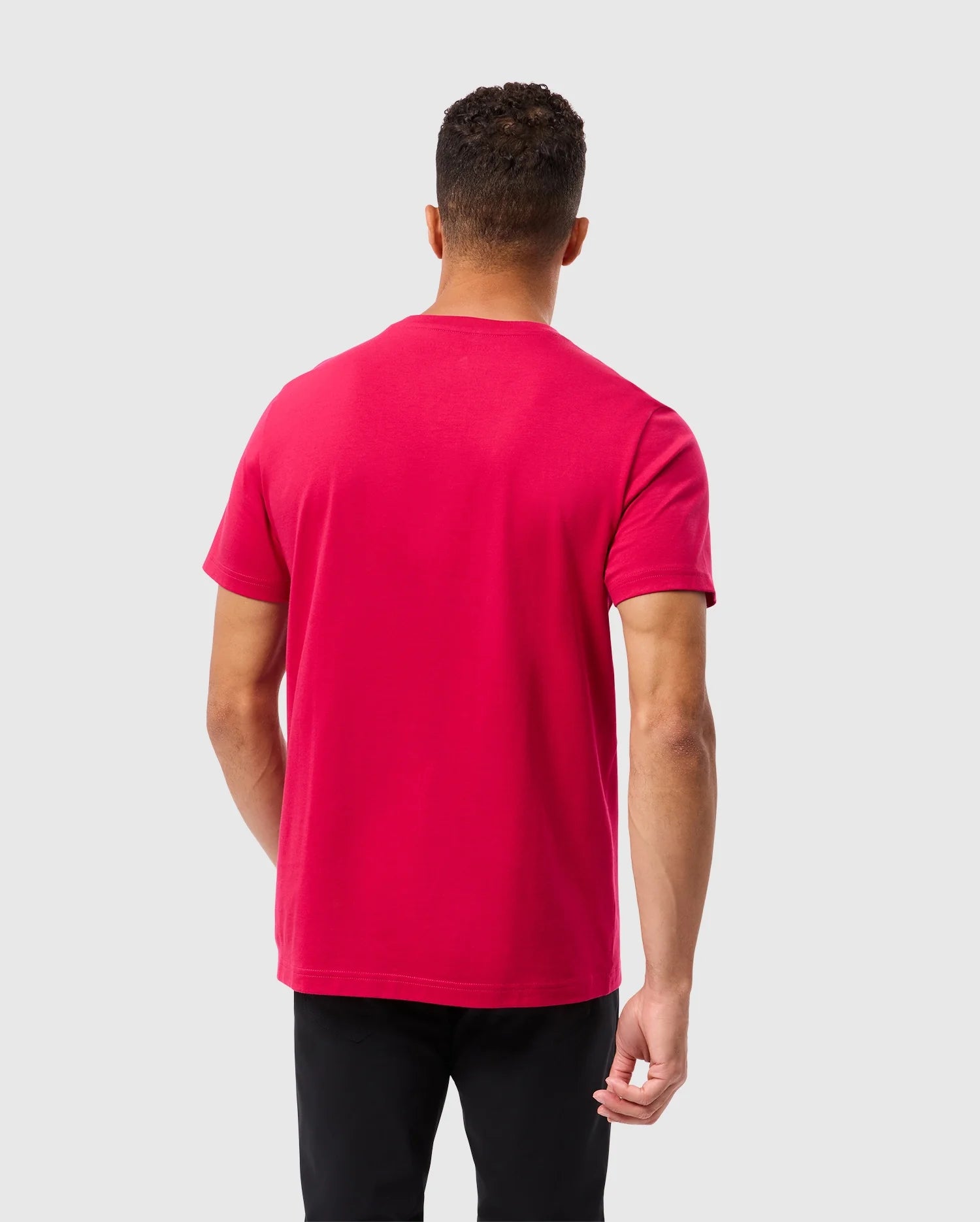 A person with short curly hair is standing with their back to the camera, wearing a vibrant red Psycho Bunny MENS TACOMA GRAPHIC TEE - B6U909D200 and black pants. The fabric of the shirt looks soft, possibly Pima cotton. The background is a plain, light grey.
