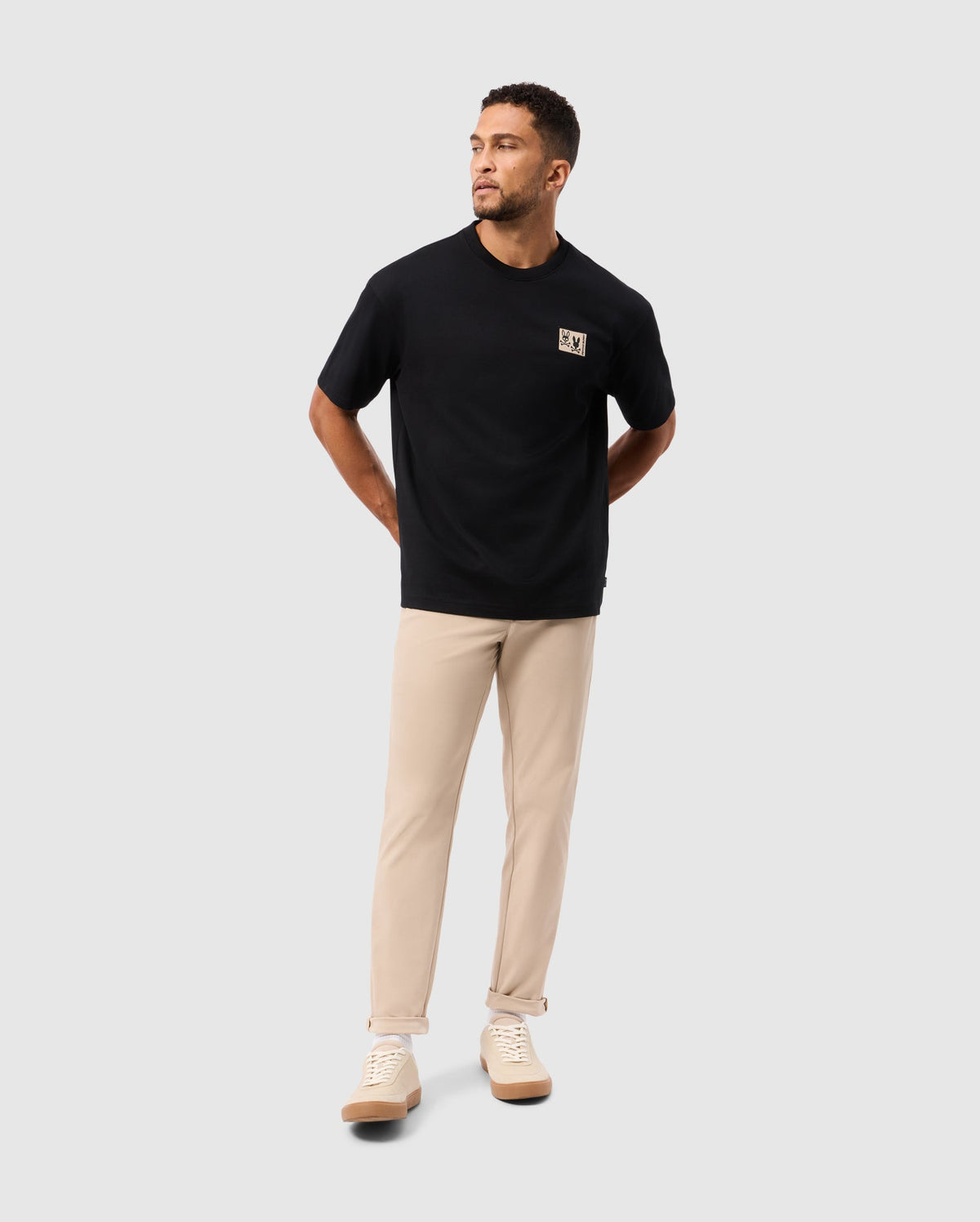 A man wearing an oversized black MENS MONROE HEAVY WEIGHT RELAXED FIT TEE by Psycho Bunny and beige pants stands against a light gray background. With his hands behind his back, he gazes to the side in a relaxed fit style, complemented by light-colored sneakers.