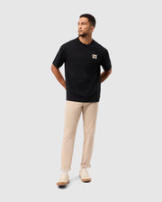 A man wearing an oversized black MENS MONROE HEAVY WEIGHT RELAXED FIT TEE by Psycho Bunny and beige pants stands against a light gray background. With his hands behind his back, he gazes to the side in a relaxed fit style, complemented by light-colored sneakers.