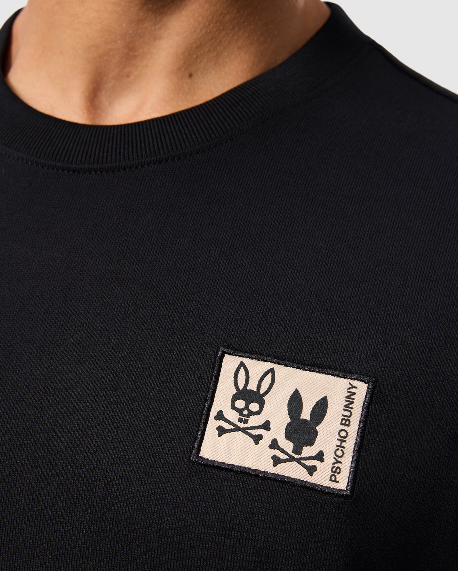 A close-up showcases a person wearing the MENS MONROE HEAVY WEIGHT RELAXED FIT TEE - B6U910D200 from Psycho Bunny. This black T-shirt, crafted from soft Pima cotton, features an oversized style with a rectangular patch on the chest. The patch displays a black bunny with crossbones and the text "Psycho Bunny" on a beige background, giving it an edgy touch.