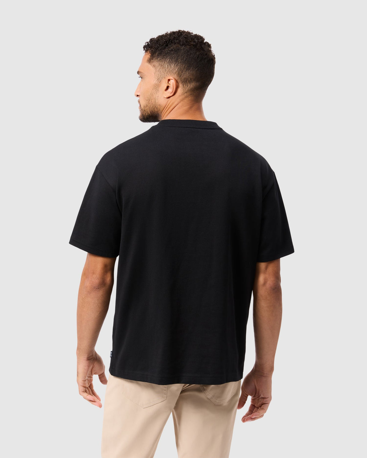 A person with short curly hair is standing and facing away from the camera. They are wearing the Psycho Bunny MENS MONROE HEAVY WEIGHT RELAXED FIT TEE in black alongside beige pants against a neutral gray background.