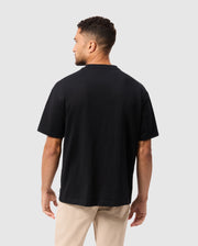 A person with short curly hair is standing and facing away from the camera. They are wearing the Psycho Bunny MENS MONROE HEAVY WEIGHT RELAXED FIT TEE in black alongside beige pants against a neutral gray background.