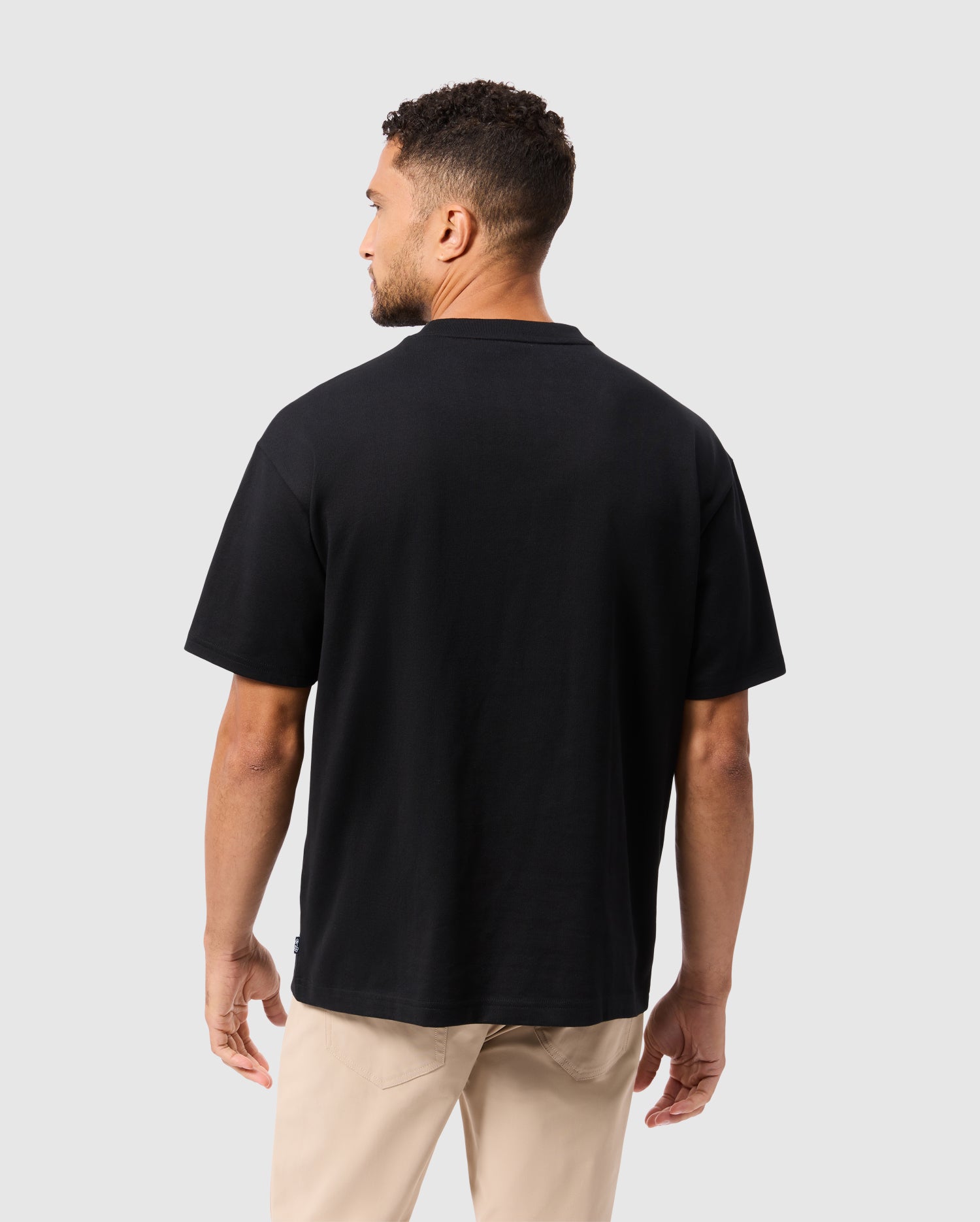 A person with short curly hair is standing and facing away from the camera. They are wearing the Psycho Bunny MENS MONROE HEAVY WEIGHT RELAXED FIT TEE in black alongside beige pants against a neutral gray background.