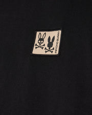 The MENS MONROE HEAVY WEIGHT RELAXED FIT TEE by Psycho Bunny features a black Pima cotton fabric with a beige patch displaying two logos: one with a skull and bunny ears, and the other a simple bunny silhouette. The patch includes the text "Psycho Bunny" printed vertically, perfectly suited for an oversized t-shirt with a relaxed fit.