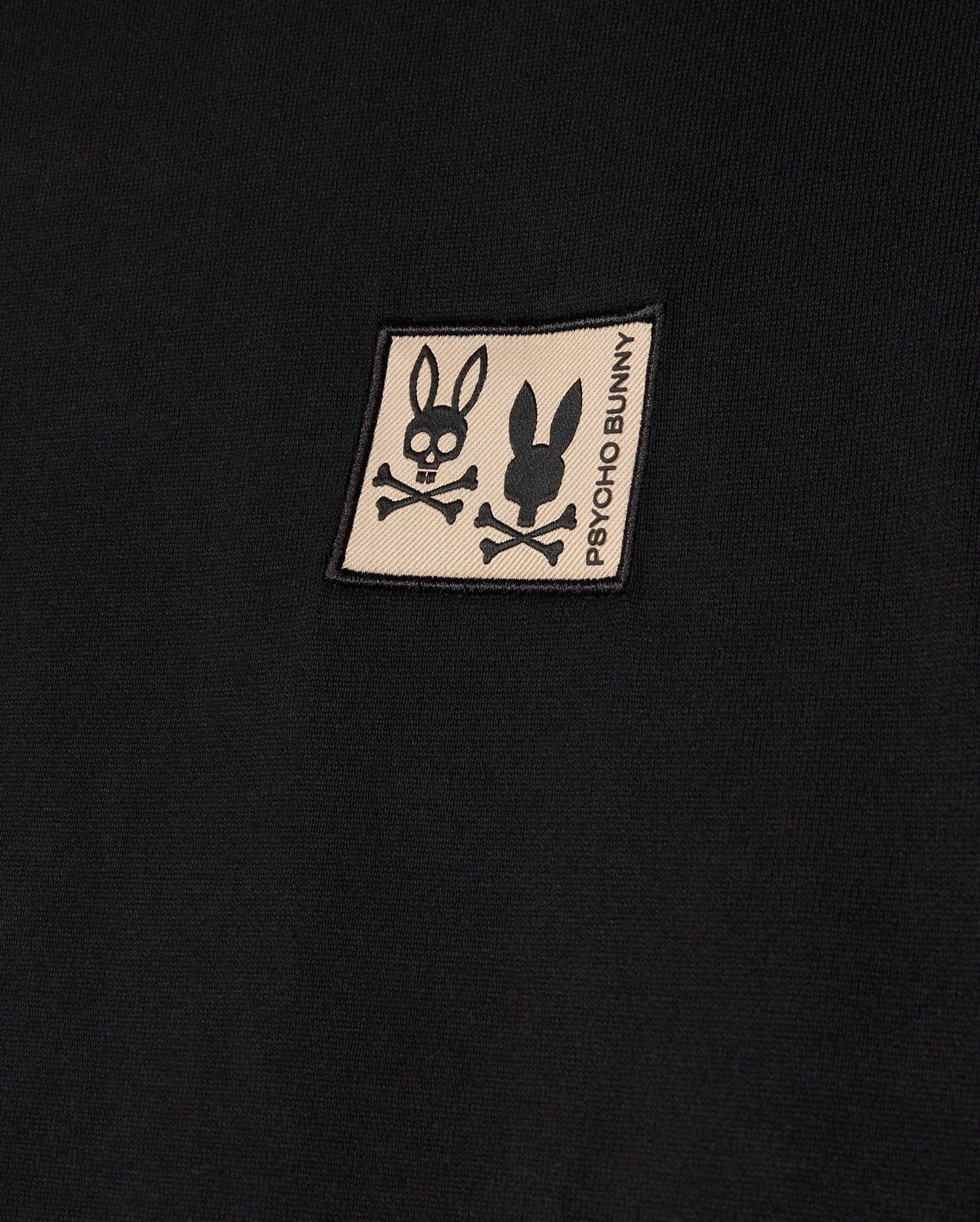 The MENS MONROE HEAVY WEIGHT RELAXED FIT TEE by Psycho Bunny features a black Pima cotton fabric with a beige patch displaying two logos: one with a skull and bunny ears, and the other a simple bunny silhouette. The patch includes the text "Psycho Bunny" printed vertically, perfectly suited for an oversized t-shirt with a relaxed fit.