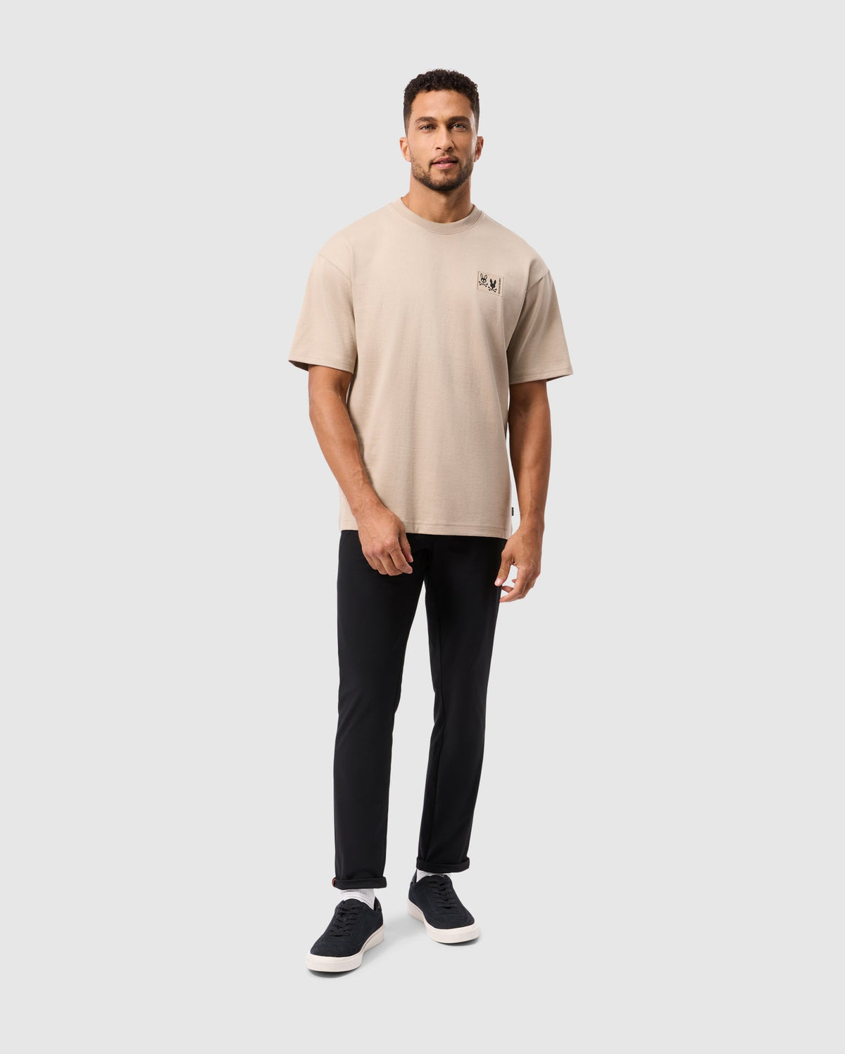 A man in a Psycho Bunny MENS MONROE HEAVY WEIGHT RELAXED FIT TEE, black pants, and sneakers exudes casual style against a plain white backdrop. The beige Pima cotton tee features a small logo on the left chest.