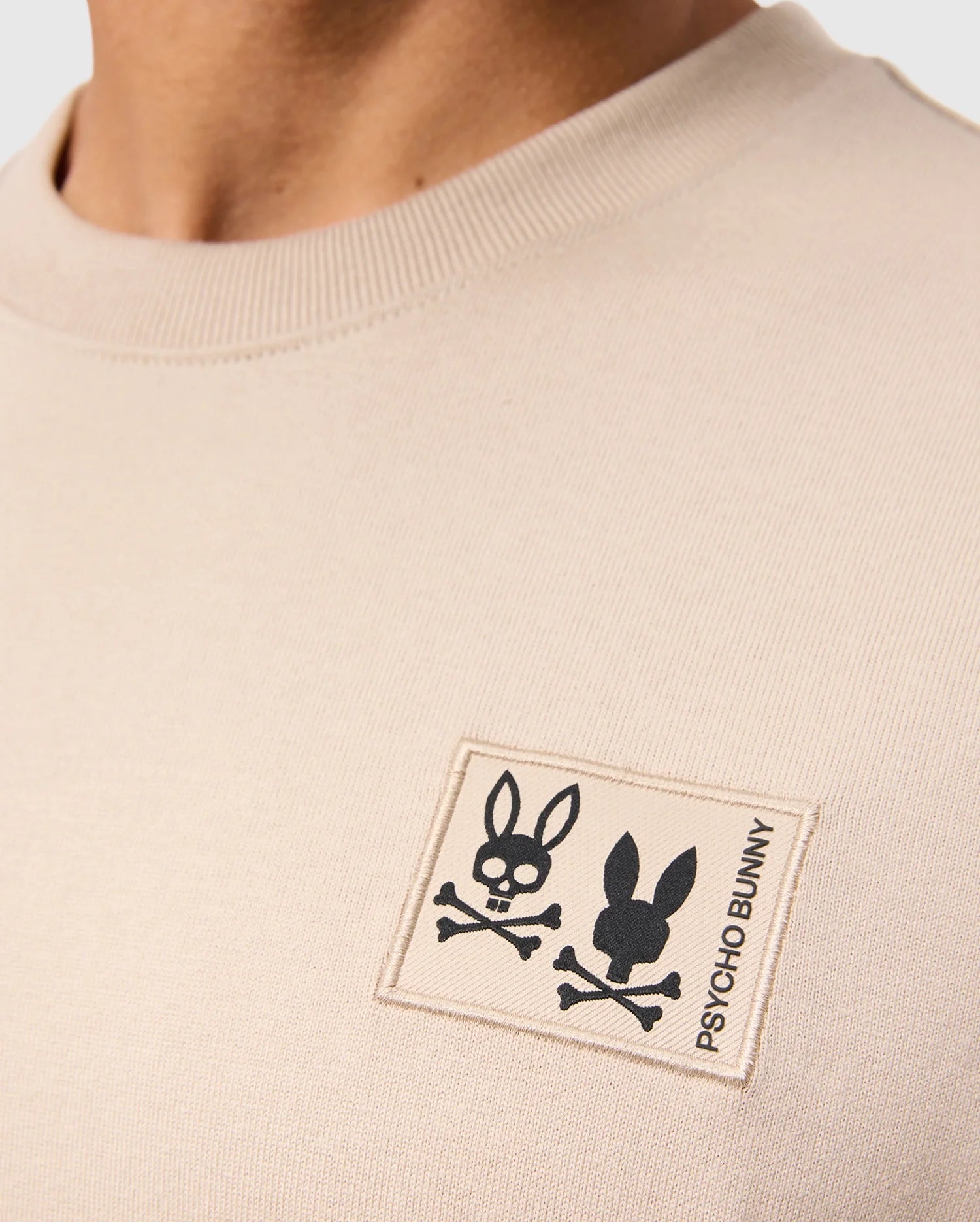 Close-up of the beige Monroe Heavy Weight Relaxed Fit Tee by Psycho Bunny. It's a casual Pima cotton shirt featuring a chest logo patch with two bunny skull and crossbones designs alongside the "Psycho Bunny" text.