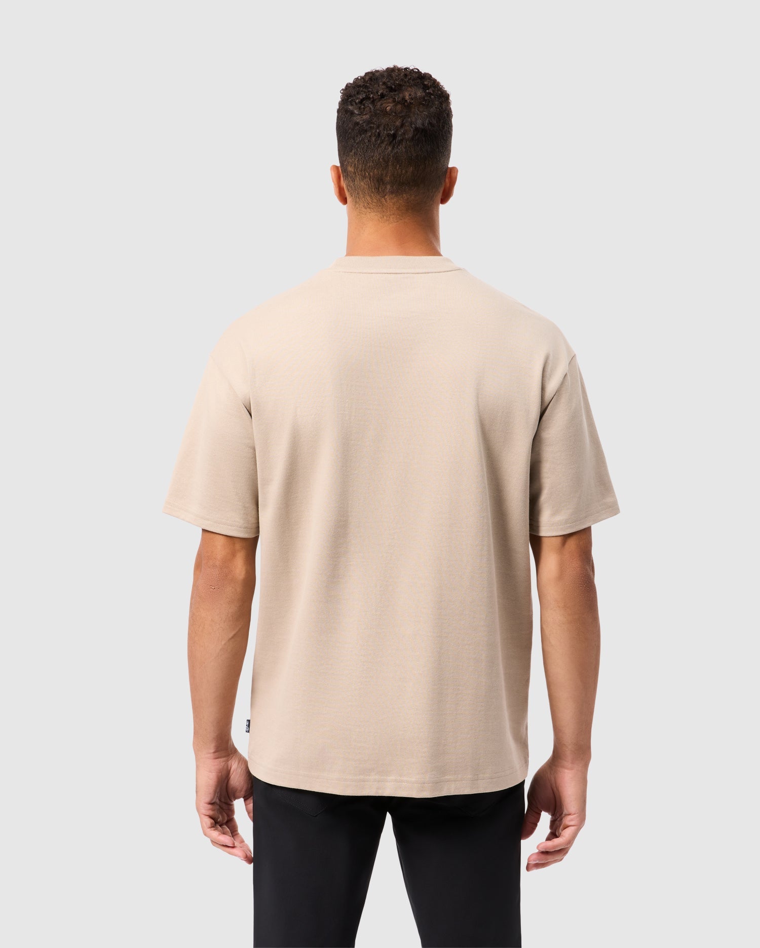A person with short curly hair wears a beige Psycho Bunny Monroe heavy weight relaxed fit tee and black pants, showcasing a casual style as they stand against a plain light gray background, facing away from the camera.