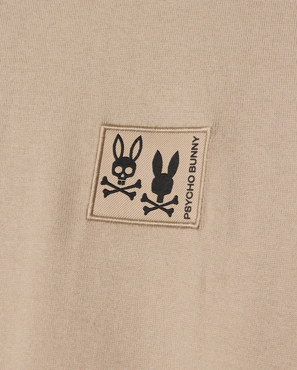 The MENS MONROE HEAVY WEIGHT RELAXED FIT TEE by Psycho Bunny highlights a close-up of its beige Pima cotton fabric, featuring a patch with two bunny skull and crossbones designs and the text "Psycho Bunny," combining edgy charm with casual elegance.