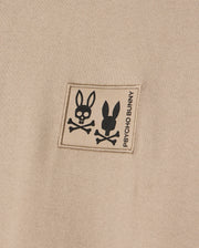 The MENS MONROE HEAVY WEIGHT RELAXED FIT TEE by Psycho Bunny highlights a close-up of its beige Pima cotton fabric, featuring a patch with two bunny skull and crossbones designs and the text "Psycho Bunny," combining edgy charm with casual elegance.