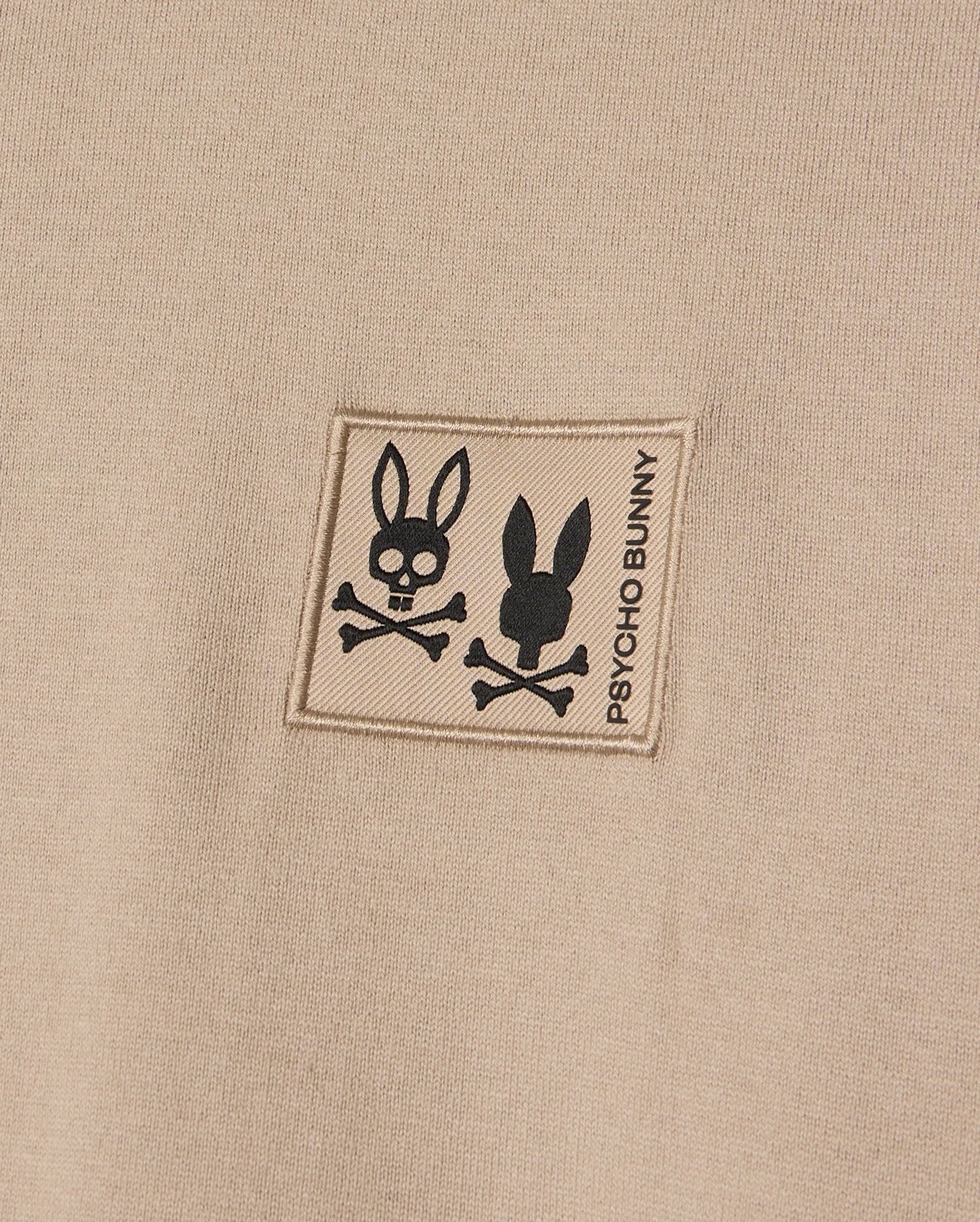The MENS MONROE HEAVY WEIGHT RELAXED FIT TEE by Psycho Bunny highlights a close-up of its beige Pima cotton fabric, featuring a patch with two bunny skull and crossbones designs and the text "Psycho Bunny," combining edgy charm with casual elegance.