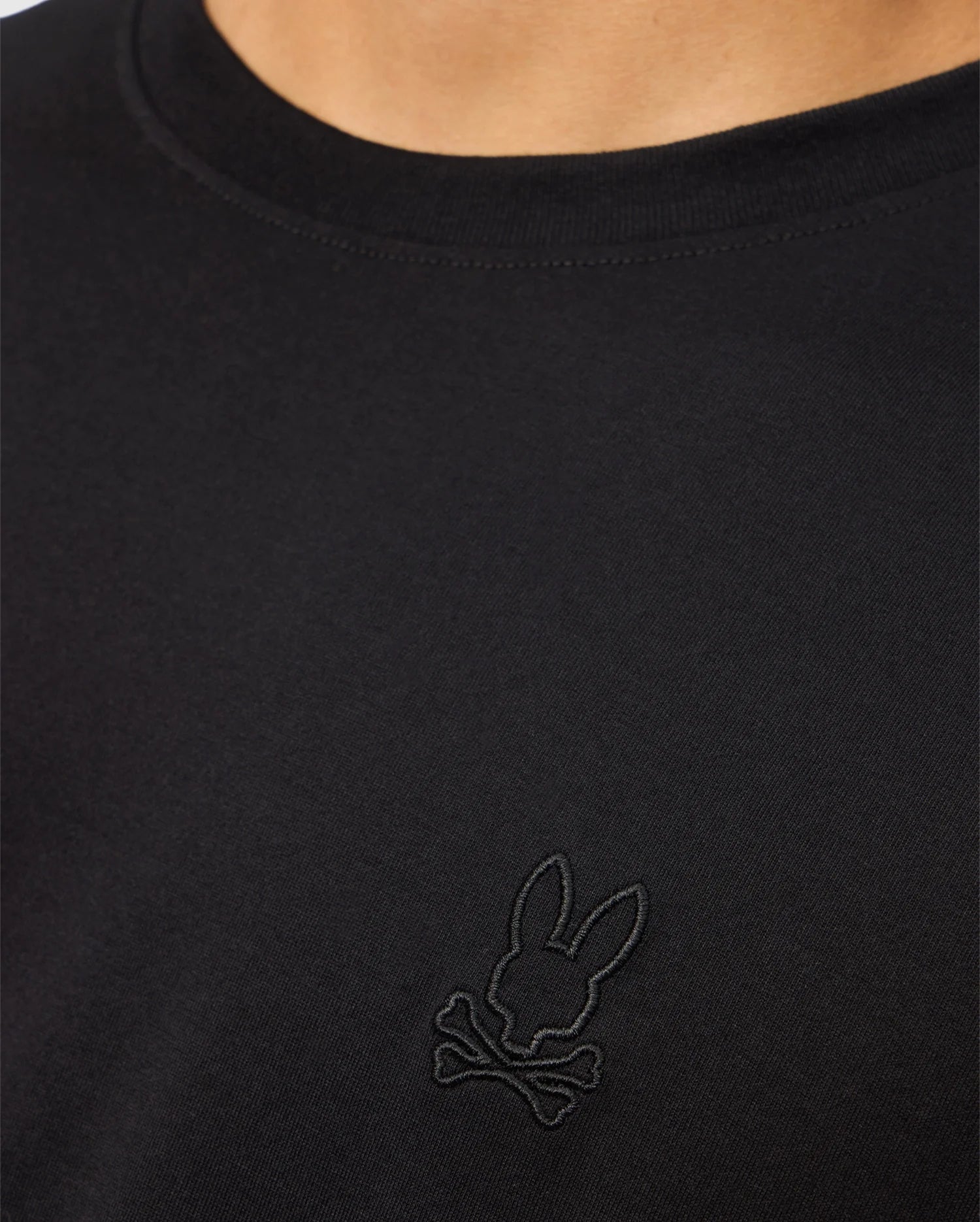 Close-up of a person wearing the Psycho Bunny MENS JAMES PREMIUM CREW NECK TEE - B6U952D200, a regular fit black T-shirt made from Premium Pima cotton, showcasing intricate embroidery of a rabbit's head above two crossed bones on the chest.