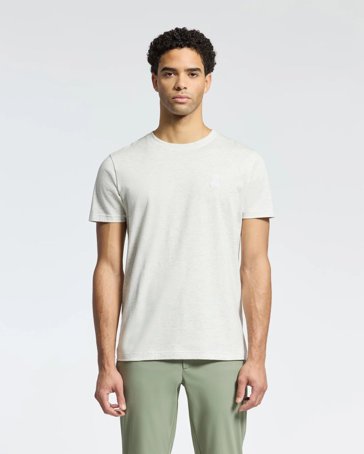 A person models the MENS JAMES CREW NECK TEE by Psycho Bunny in light gray Pima cotton paired with olive green pants against a plain backdrop, highlighting exquisite craftsmanship with subtle embroidery, all while maintaining a neutral expression.