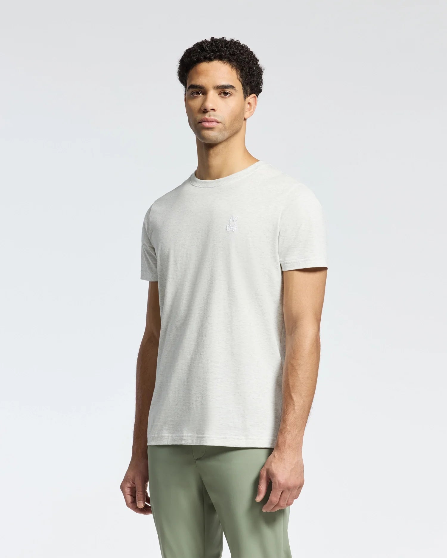 A person wearing a Psycho Bunny MEN'S JAMES CREW NECK TEE - B6U952D200 in light gray stands against a plain background. The tee showcases fine craftsmanship with a small, subtle chest design. They pair it with light green pants and have short, curly hair.