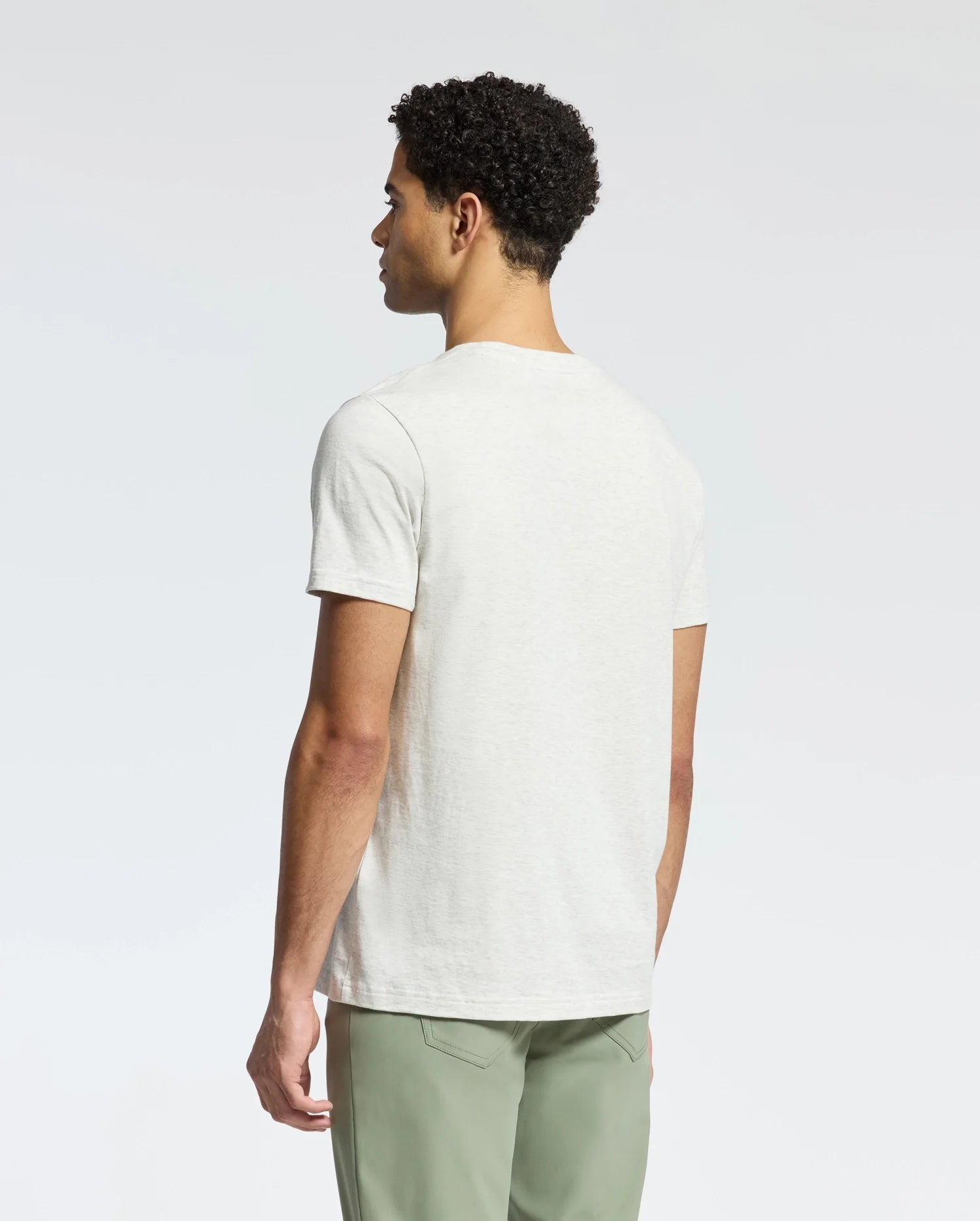 A person with short curly hair is seen from behind, wearing a plain white Psycho Bunny MEN'S JAMES CREW NECK TEE and light green pants against a simple light gray background.