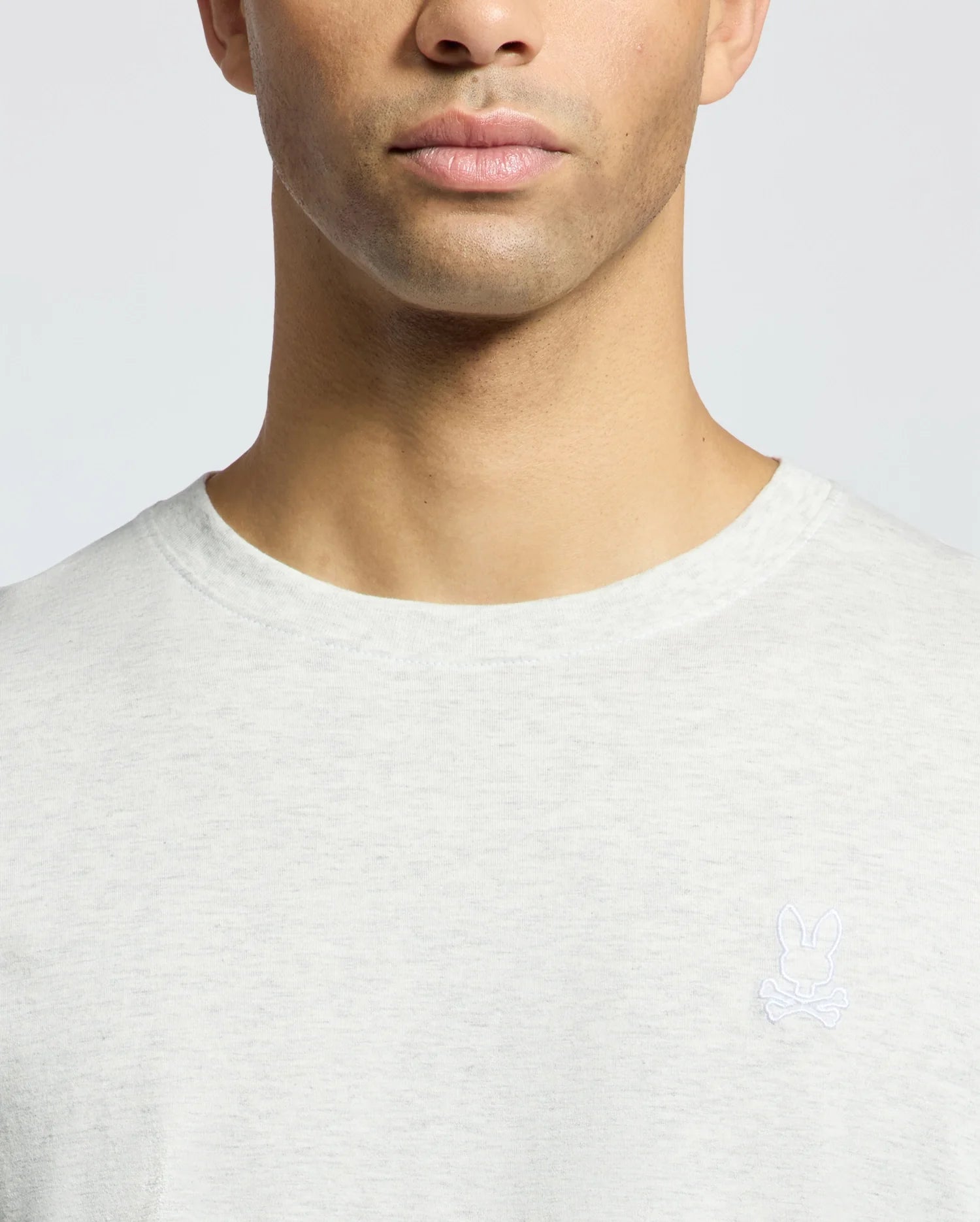 A person wears the Psycho Bunny Mens James Crew Neck Tee (B6U952D200) in light grey, showcasing a small embroidered rabbit and crossbones logo on the chest. The image captures the upper torso and neck against a plain background, emphasizing its fine craftsmanship.
