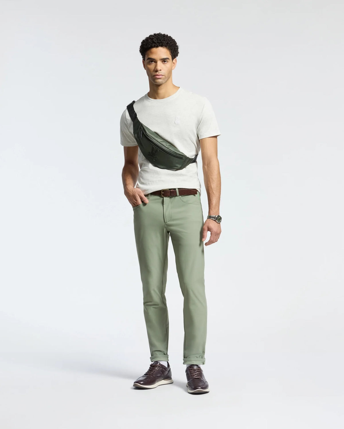 A man stands against a plain background wearing a Psycho Bunny MENS JAMES CREW NECK TEE in white, green pants, and brown shoes. He accessorizes with a black crossbody bag, brown belt, and displays craftsmanship with a watch on his left wrist.