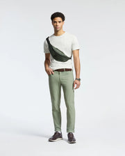 A man stands against a plain background wearing a Psycho Bunny MENS JAMES CREW NECK TEE in white, green pants, and brown shoes. He accessorizes with a black crossbody bag, brown belt, and displays craftsmanship with a watch on his left wrist.
