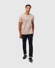 A man stands against a plain white background, wearing a beige Psycho Bunny MENS JAMES PREMIUM CREW NECK TEE - B6U952D200 made from premium Pima cotton and black pants. He is also wearing black sneakers. He has short, curly hair and is smiling slightly while looking towards the camera.
