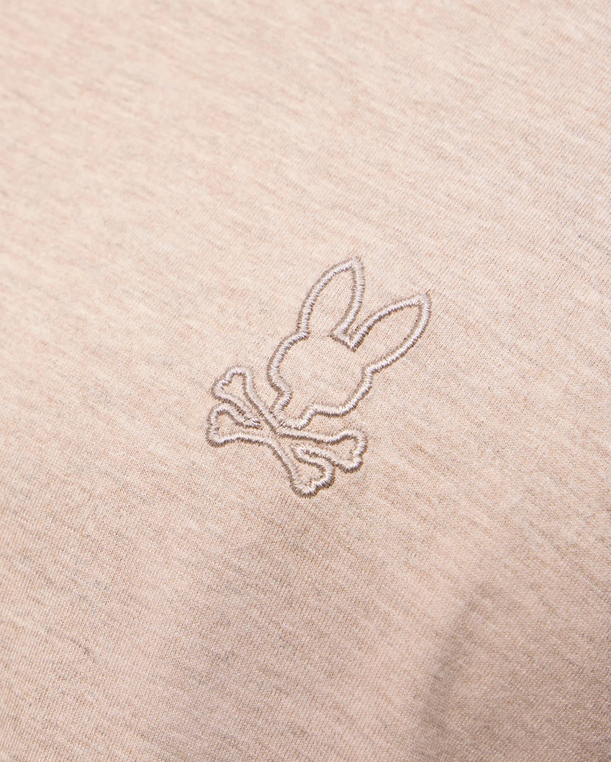 Close-up photo of beige fabric displaying an embroidered design on the Psycho Bunny MENS JAMES PREMIUM CREW NECK TEE - B6U952D200. The meticulous embroidery features a stylized bunny head above a pair of crossed bones, reminiscent of a skull and crossbones motif. The stitching is in a slightly darker shade than the premium Pima cotton fabric.