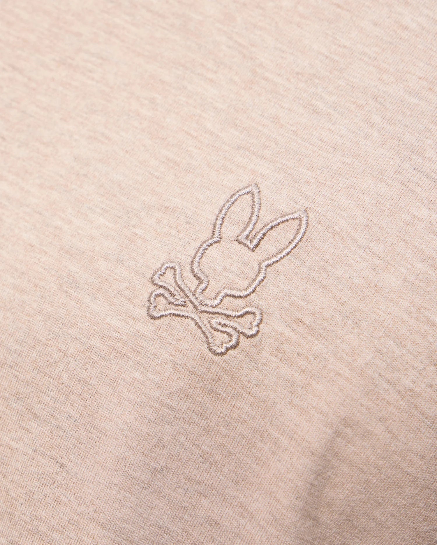 Close-up photo of beige fabric displaying an embroidered design on the Psycho Bunny MENS JAMES PREMIUM CREW NECK TEE - B6U952D200. The meticulous embroidery features a stylized bunny head above a pair of crossed bones, reminiscent of a skull and crossbones motif. The stitching is in a slightly darker shade than the premium Pima cotton fabric.