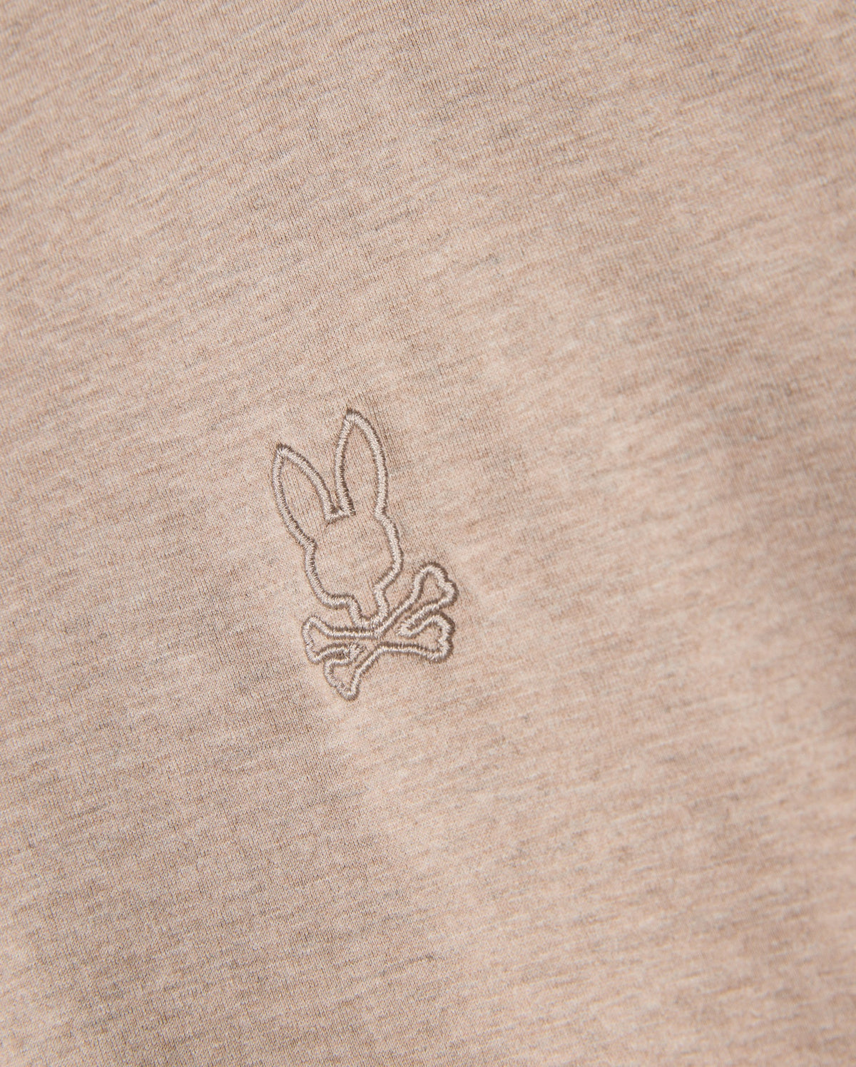 Close-up of a beige Psycho Bunny MENS JAMES PREMIUM CREW NECK TEE - B6U952D200, crafted from premium Pima cotton. The tee features a small, meticulously embroidered design of a bunny head above two crossed bones, resembling a playful variation of the skull and crossbones symbol. The stitching is in a slightly darker shade than the fabric.
