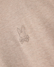 Close-up of a beige Psycho Bunny MENS JAMES PREMIUM CREW NECK TEE - B6U952D200, crafted from premium Pima cotton. The tee features a small, meticulously embroidered design of a bunny head above two crossed bones, resembling a playful variation of the skull and crossbones symbol. The stitching is in a slightly darker shade than the fabric.
