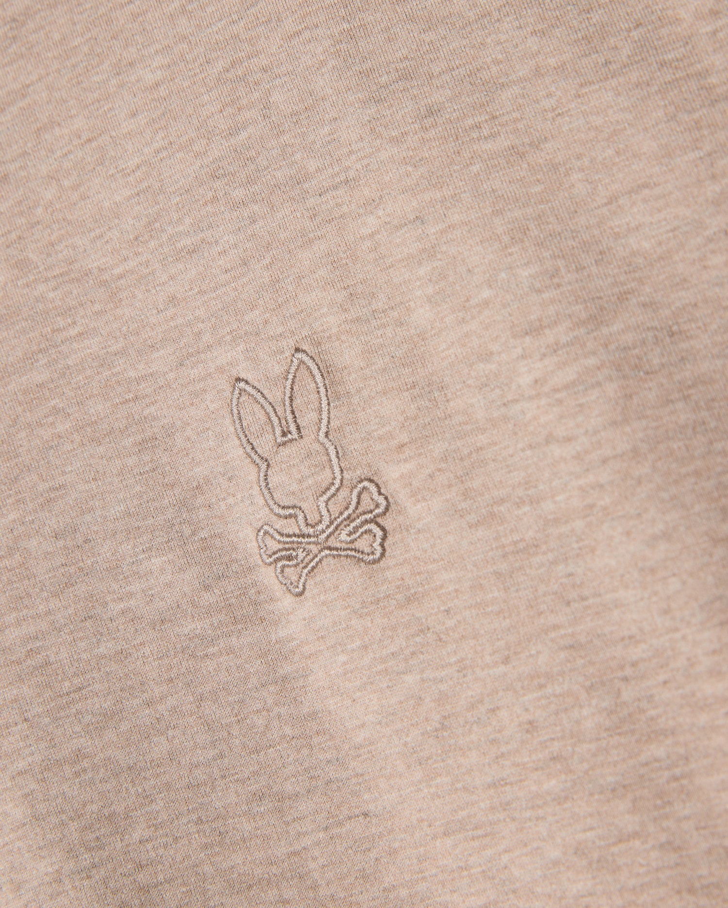 Close-up of a beige Psycho Bunny MENS JAMES PREMIUM CREW NECK TEE - B6U952D200, crafted from premium Pima cotton. The tee features a small, meticulously embroidered design of a bunny head above two crossed bones, resembling a playful variation of the skull and crossbones symbol. The stitching is in a slightly darker shade than the fabric.
