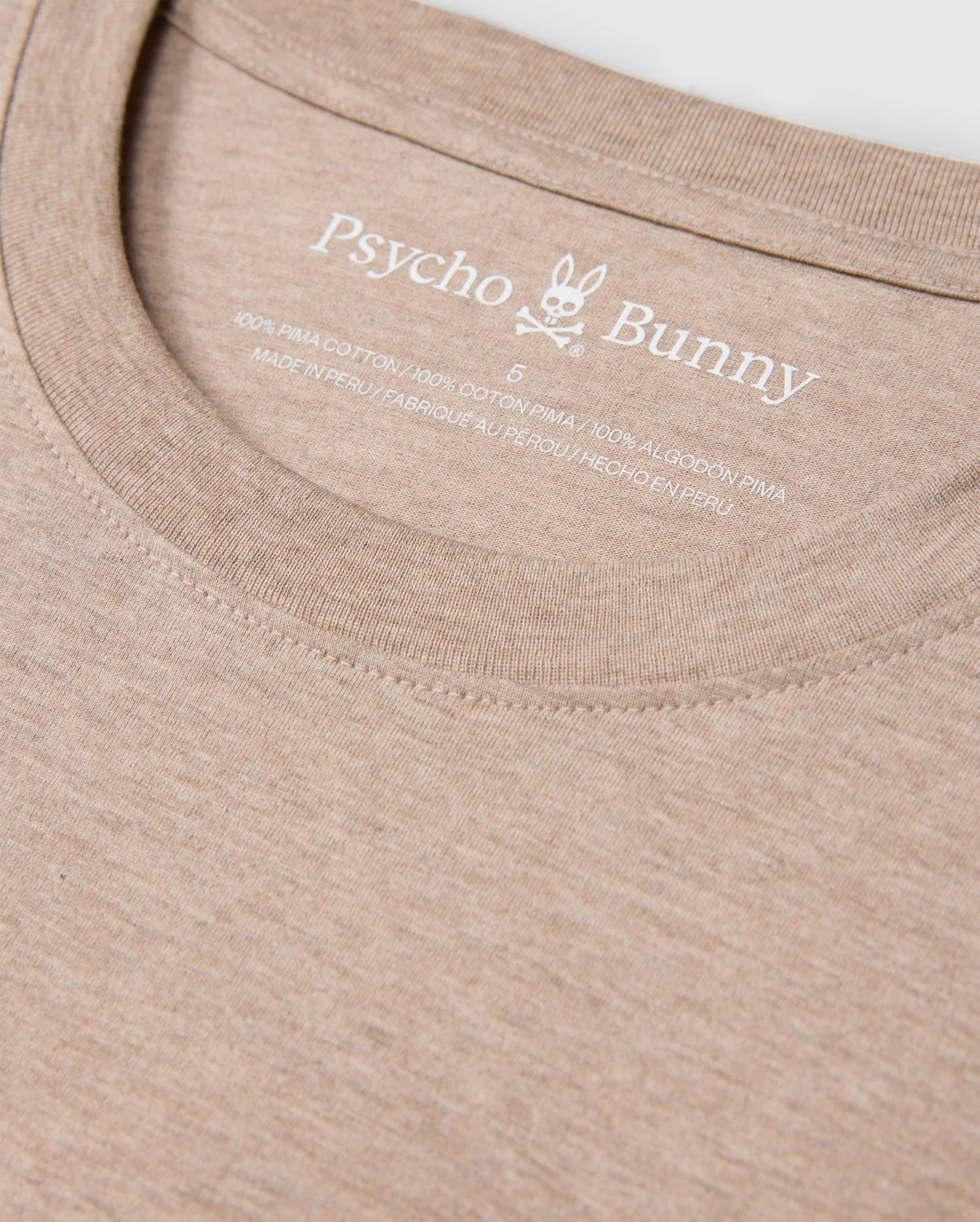 Close-up of the beige collar on the Psycho Bunny MENS JAMES PREMIUM CREW NECK TEE - B6U952D200, featuring a printed label with a small bunny skull and crossbones logo. The label indicates that this premium Pima cotton shirt is size 6 and crafted in Peru.